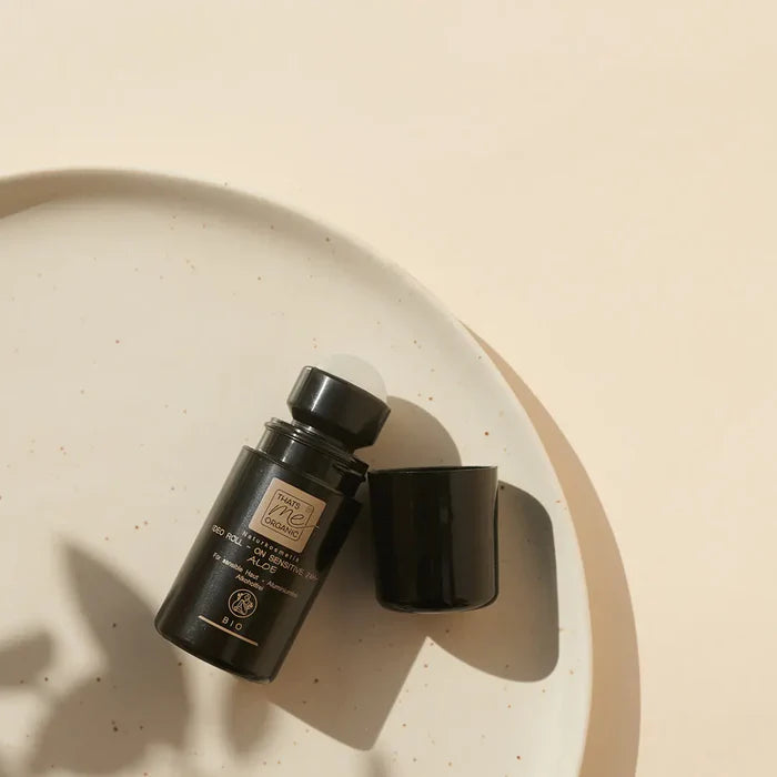 Geöffneter schwarzer „THATS me ORGANIC“ Deo Roll-on mit sichtbarer Kugel und danebenliegender Kappe auf einem hellen, gesprenkelten Keramikteller bei hellem Sonnenlicht.