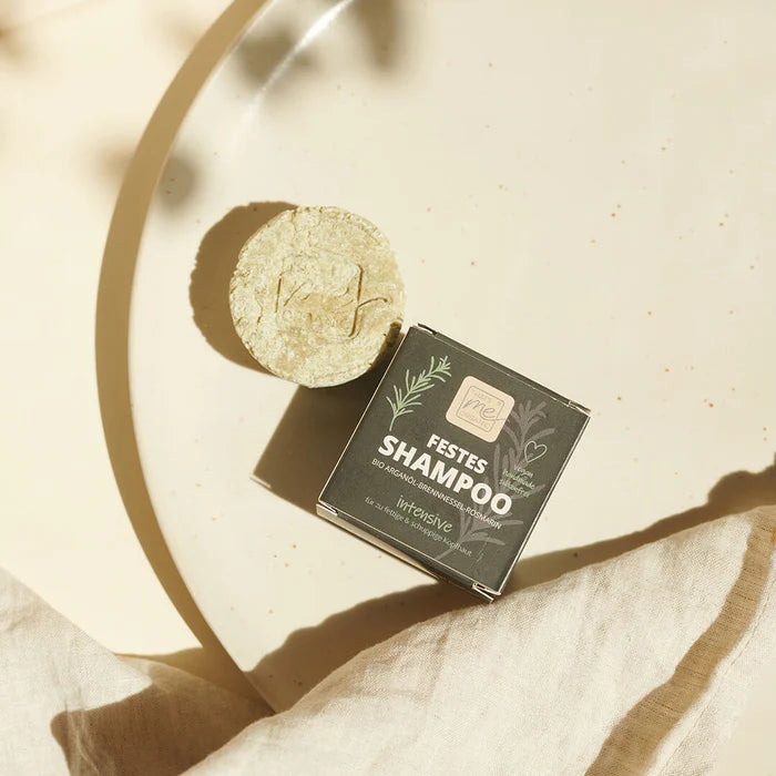 Minimalistische Produktfotografie bei idery: Ein rundes, hellgrünes festes Shampoo-Stück liegt neben seiner quadratischen, schwarzen Verpackung mit der Aufschrift 'FESTES SHAMPOO intensive'. Die Produkte sind dekorativ auf einem großen, cremefarbenen Keramikteller platziert, der teilweise von einem hellen Leinentuch bedeckt ist. Sanftes Sonnenlicht wirft dezente Blätterschatten über die gesamte Szene.