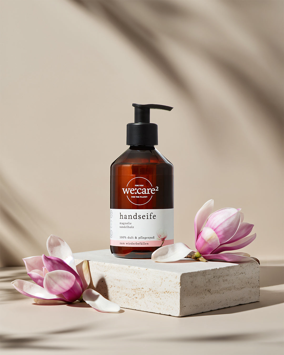 Ästhetische Produktpräsentation von Handseife bei idery (we:care²): Die braune Flasche mit Magnolien-Duft steht auf einem hellen Natursteinsockel, umrahmt von zarten Magnolienblüten. Ein Spiel aus Licht und Schatten im Hintergrund unterstreicht das hochwertige, nachhaltige Design der Pflegeprodukte.
